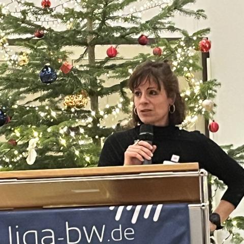 Junge Frau steht hinter Stehpult mit Mikrophon in der Hand. Hinter ihr ist ein Weihnachtsbaum zu sehen 