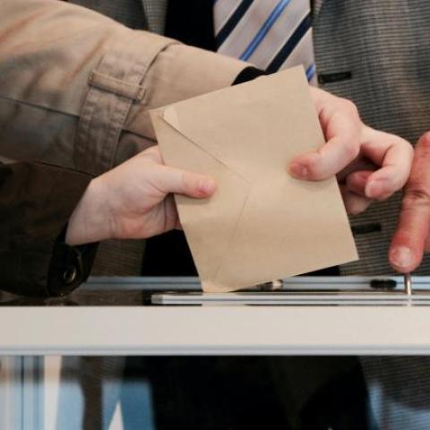 eine Hand steckt einen Umschlag in eine Wahlurne