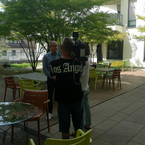 Kameramann und Reporterin im Innenhof mit Vorstand vor Bäumen