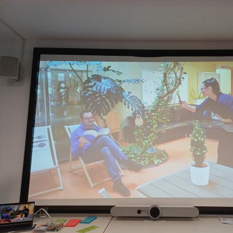 Auf einem Bildschirm wird eine Szene gezeigt, bei der eine Frau ein Foto von einem sitzenden Mann macht.