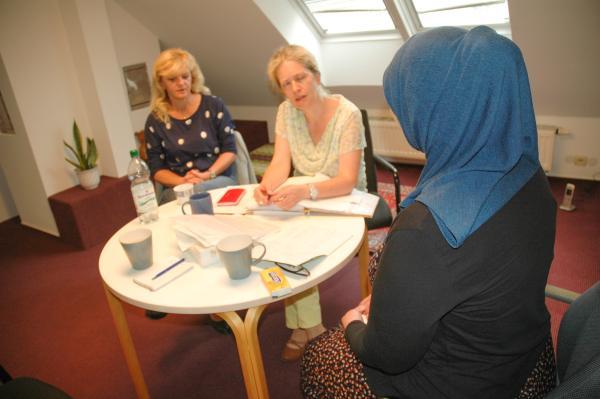 zwei Frauen an einem kleinen runden Tisch gegenüber eine Frau im blauen Kopftuch von hinten