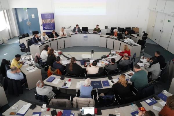 große Beratungsrunde mit vielen Menschen an im Kreis platzierten Tischen