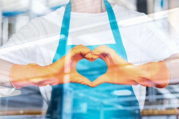 Person mit blauer Schürze und gelben Handschuhen formt ein Herz mit den Händen