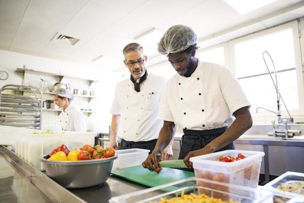 ein junger Mann in Koch-Outfit legt eine Erdbeere auf ein Backblech, ein Anleiter steht neben ihm