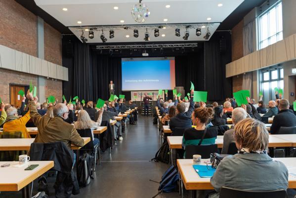 Ein Tagungssaal mit Blick auf eine Bühne, die Tagenden heben grüne Stimmzettel in die Höhe. 