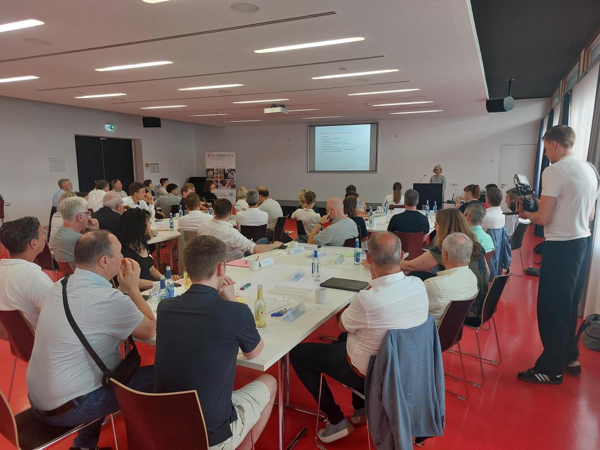 Ca. 50 Personen an Diskussionstischen, im Hintergrund steht eine Person am Rednerpult