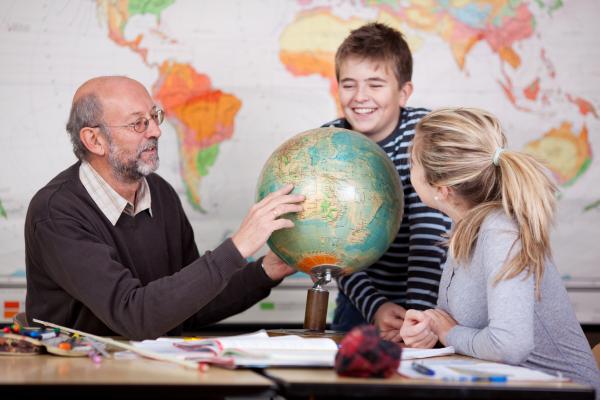 Ein sitzender Mann mit Glatze und grauem Haar erklärt im Klassenzimmer einem dunkelhaarigen Jungen und einem blonden Mädchen im Alter von 10 Jahren die Weltkugel