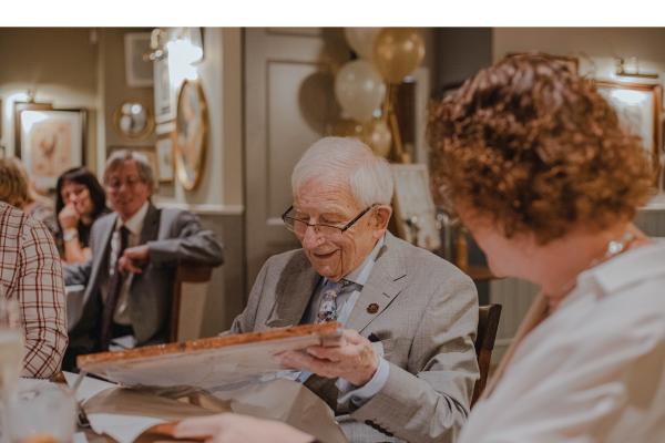 Älterer Mann sitzt am Tisch und hält Stück von Spanplatte in der hand