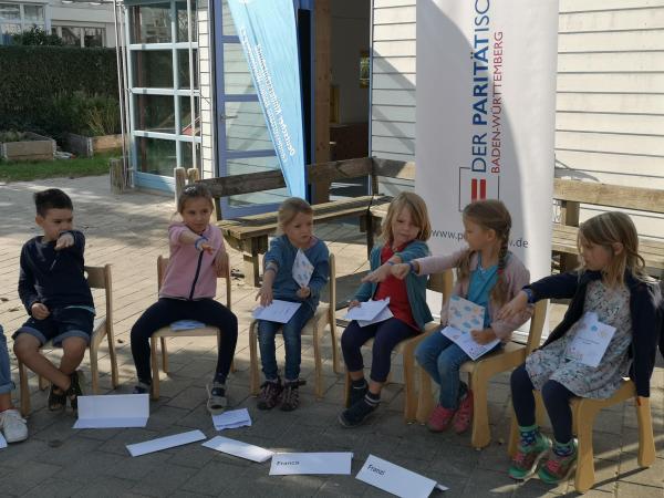 Kleine Mädchen und Jungs sitzen im Stuhlkreis und strechen ihren Arm mit dem Dankeschönarmbändchen raus