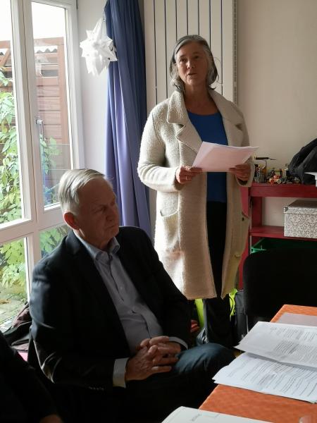 Sitzender Mann in dunklem Anzug und stehende Frau mit weißer langer Jacke