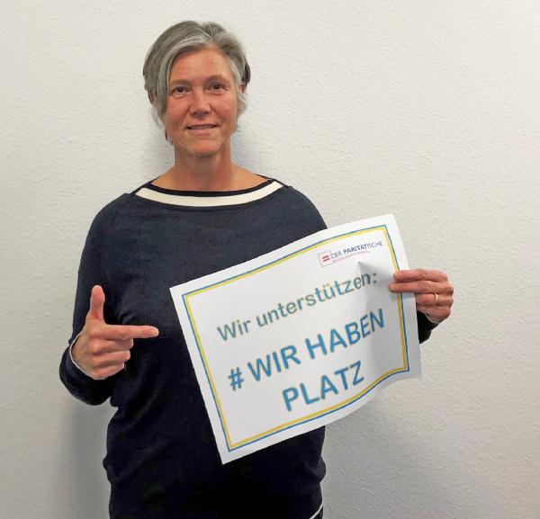 Stehende Frau hält ein Plakat in einer Hand und deutet mit der anderen auf die Aufschrift #Wir haben Platz